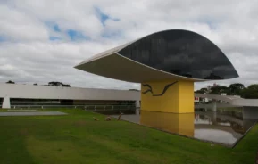 Um edifício moderno com um grande telhado preto em formato oval apoiado em uma base amarela, cercado por grama e água, sob um céu nublado.