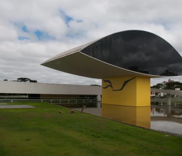 Um edifício moderno com um grande telhado preto em formato oval apoiado em uma base amarela, cercado por grama e água, sob um céu nublado.