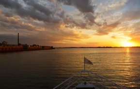 Um barco segue em direção a um pôr do sol dourado em águas calmas, com armazéns e uma chaminé de fumaça ao longo da costa esquerda sob um céu parcialmente nublado. A imagem mostra o pôr do sol no Guaíba, um dos pontos turísticos de Porto Alegre.