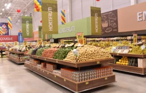 A seção de hortifrutigranjeiros de um supermercado bem iluminado exibe frutas e legumes bem organizados, incluindo batatas, cebolas e bananas. Balões coloridos estão pendurados no teto e as placas acima indicam HORTIFRUTI.