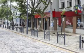 Imagem da Alameda Prudente de Moraes, em Curitiba, mostra uma rua de paralelepípedos ladeada por grades de metal, árvores e lojas, incluindo um prédio de fachada vermelha com bandeiras da França e do Brasil. A rua parece vazia e calma durante o dia.