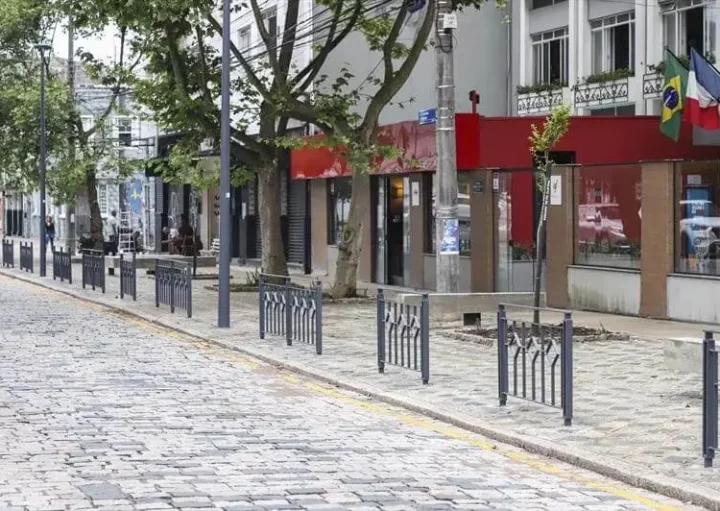 Imagem da Alameda Prudente de Moraes, em Curitiba, mostra uma rua de paralelepípedos ladeada por grades de metal, árvores e lojas, incluindo um prédio de fachada vermelha com bandeiras da França e do Brasil. A rua parece vazia e calma durante o dia.