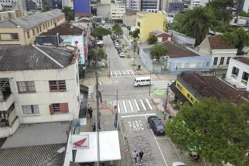 Imagem da vista aérea da Avenida Prudente de Moraes, em Curitiba, mostra um cruzamento urbano com ruas de paralelepípedos, faixas de pedestres, carros estacionados, árvores e edifícios em várias cores, incluindo amarelo e azul.