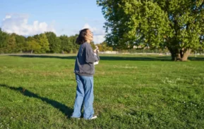 Imagem de uma mulher vestindo uma jaqueta cinza e calça jeans azul na grama verde de um parque ensolarado, olhando para cima e sorrindo, com árvores e um céu azul ao fundo, para ilustrar matéria sobre o bairro mais arborizado de SP.