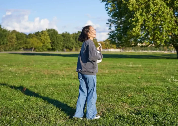 Imagem de uma mulher vestindo uma jaqueta cinza e calça jeans azul na grama verde de um parque ensolarado, olhando para cima e sorrindo, com árvores e um céu azul ao fundo, para ilustrar matéria sobre o bairro mais arborizado de SP.