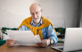 Imagem de um homem mais velho, de óculos e com um suéter amarelo sobre os ombros, senta-se à mesa, lendo papéis, com um laptop aberto à sua frente em um ambiente doméstico.