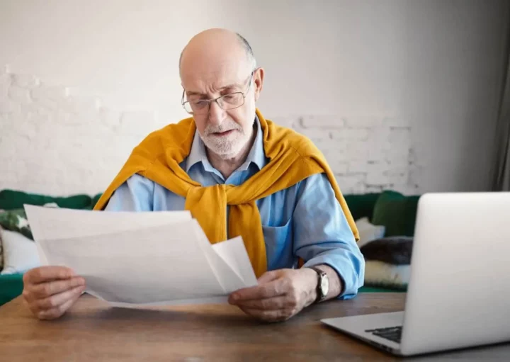 Imagem de um homem mais velho, de óculos e com um suéter amarelo sobre os ombros, senta-se à mesa, lendo papéis, com um laptop aberto à sua frente em um ambiente doméstico.