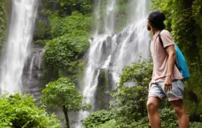 Imagem de uma pessoa com roupas casuais e boné em frente a uma floresta verde, olhando para uma alta cachoeira que desce em cascata pelas rochas ao fundo.