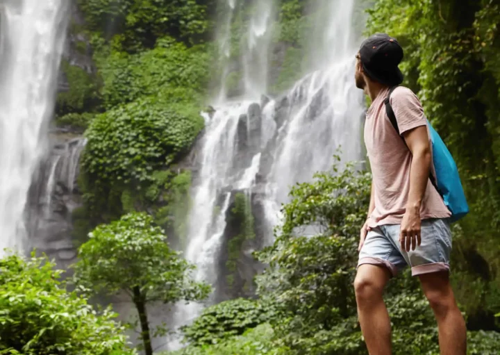 Imagem de uma pessoa com roupas casuais e boné em frente a uma floresta verde, olhando para uma alta cachoeira que desce em cascata pelas rochas ao fundo.