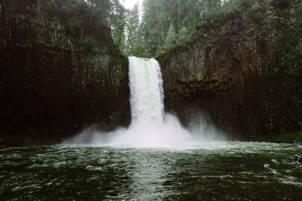 Imagem de uma poderosa cachoeira caindo em cascata sobre um penhasco rochoso cercado por uma floresta verde exuberante, criando uma névoa ao cair no rio abaixo.