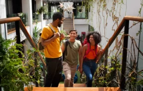 Imagem de três jovens adultos com mochilas subindo uma escada de madeira em um espaço interno iluminado e cheio de plantas, sorrindo e conversando. A área é decorada com plantas suspensas e luzes.