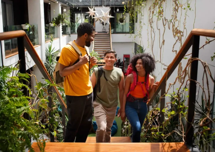 Imagem de três jovens adultos com mochilas subindo uma escada de madeira em um espaço interno iluminado e cheio de plantas, sorrindo e conversando. A área é decorada com plantas suspensas e luzes.