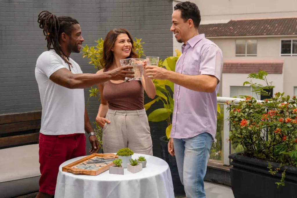 Imagem de três pessoas em um pátio, sorrindo e brindando ao redor de uma pequena mesa redonda com plantas e um jogo de tabuleiro. Plantas altas e flores decoram o ambiente externo.