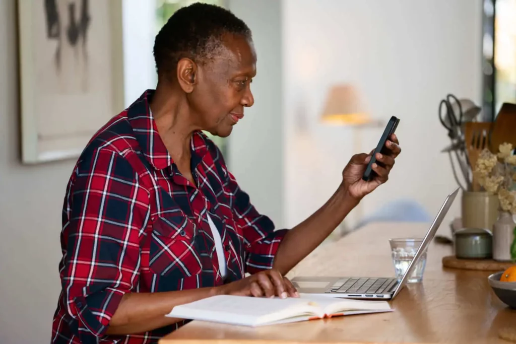 Imagem de uma mulher idosa com camisa xadrez sentada em uma mesa usando um laptop e segurando um smartphone. Um caderno aberto e um copo de água estão próximos. Ela parece concentrada em seu telefone.