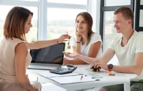 Imagem de uma mulher entregando as chaves da casa a um casal sorridente sentado em uma mesa com documentos, um laptop e uma calculadora para ilustrar matéria sobre financiamento imobiliário.