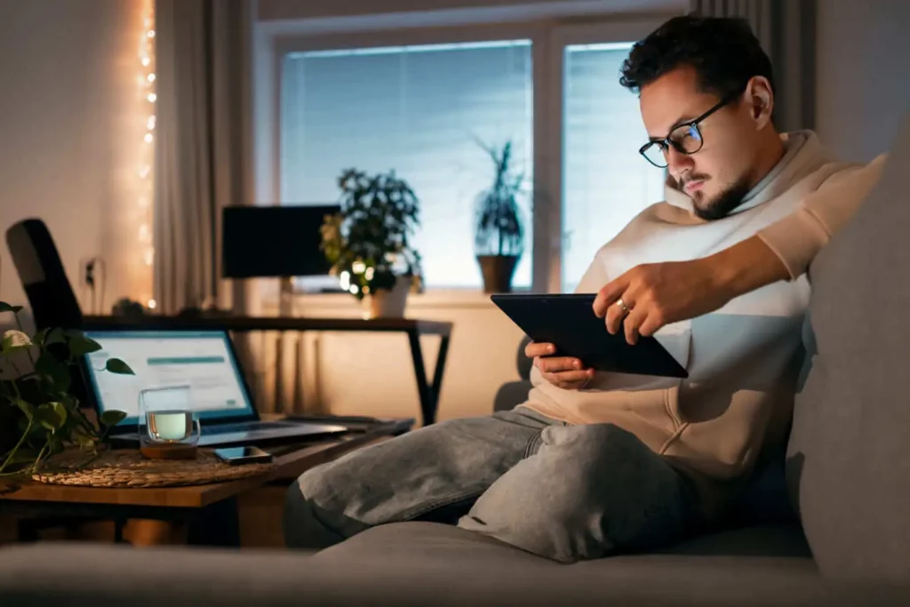 Imagem de um homem de óculos e capuz sentado em um sofá usando um tablet. Um laptop aberto, uma xícara de chá e um bloco de notas estão sobre a mesa de centro próxima. A sala tem iluminação quente, plantas e uma grande janela ao fundo.