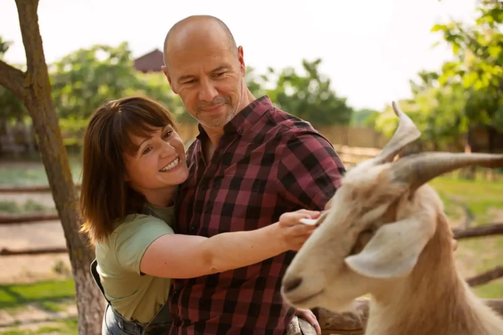 Imagem de uma mulher sorridente abraçando um homem de camisa xadrez que está acariciando uma cabra ao ar livre em uma fazenda ensolarada e arborizada. O homem olha para a cabra, e o fundo é verde e tranquilo.