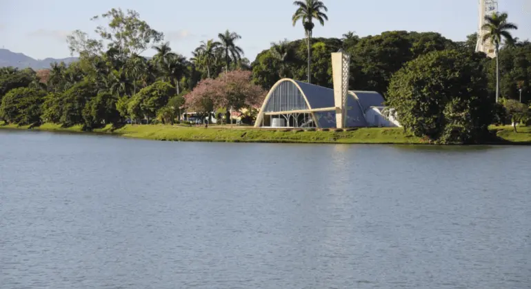 Imagem da igreja de São Francisco de Assis, na Lagoa da Pampulha, mostra a capela modernista com telhado curvo à beira do lago, cercada por árvores e vegetação sob um céu claro.
