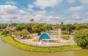 Imagem da vista aérea da Lagoa da Pampulha, em Belo Horizonte, mostra parque com uma igreja modernista azul e branca, árvores, trilhas para caminhada e um lago em primeiro plano sob um céu parcialmente nublado.