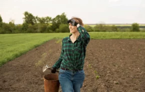 Imagem de uma mulher sorridente vestindo camisa xadrez e calça jeans em um campo, segurando um balde de metal e uma ferramenta manual, com uma mão protegendo os olhos enquanto olha para longe em um dia ensolarado.
