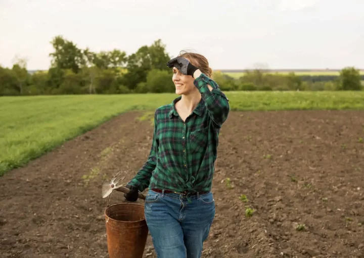 Imagem de uma mulher sorridente vestindo camisa xadrez e calça jeans em um campo, segurando um balde de metal e uma ferramenta manual, com uma mão protegendo os olhos enquanto olha para longe em um dia ensolarado.