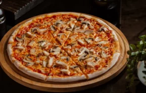 Imagem de uma pizza inteira coberta com cogumelos, queijo e molho de tomate fatiada em uma tábua de madeira, cercada por vasos de plantas e moinhos de pimenta em uma mesa escura.