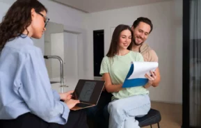 Imagem de um casal sorridente revisando documentos juntos, sentado em um escritório moderno, enquanto uma mulher com um laptop está sentada em frente a eles, possivelmente discutindo uma papelada sobre as novas regras do Minha Casa, Minha Vida.