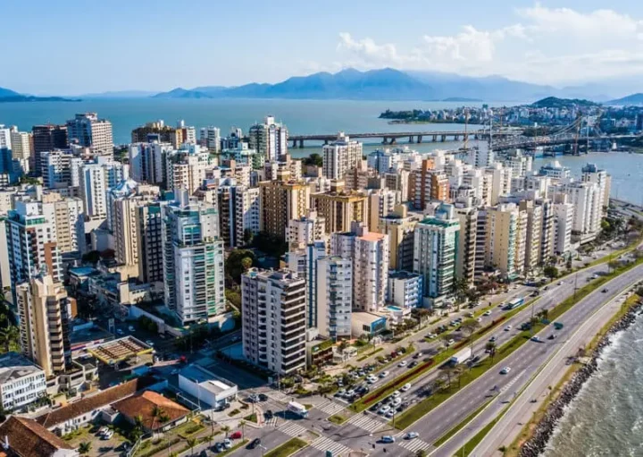 Imagem da vista aérea de Florianópolis mostra edifícios altos, uma estrada movimentada ao longo da costa e uma ponte sobre o mar; montanhas e céu azul são visíveis ao fundo.