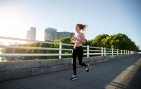 Imagem de uma mulher em trajes esportivos correndo ao longo de um caminho pavimentado em uma ponte, com prédios e árvores da cidade ao fundo, sob um céu claro e ensolarado, para ilustrar matéria sobre onde correr em SP.