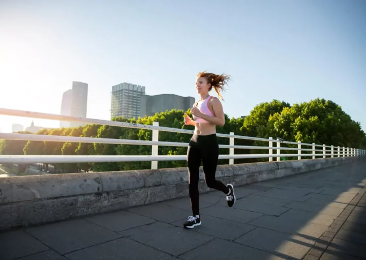 Imagem de uma mulher em trajes esportivos correndo ao longo de um caminho pavimentado em uma ponte, com prédios e árvores da cidade ao fundo, sob um céu claro e ensolarado, para ilustrar matéria sobre onde correr em SP.