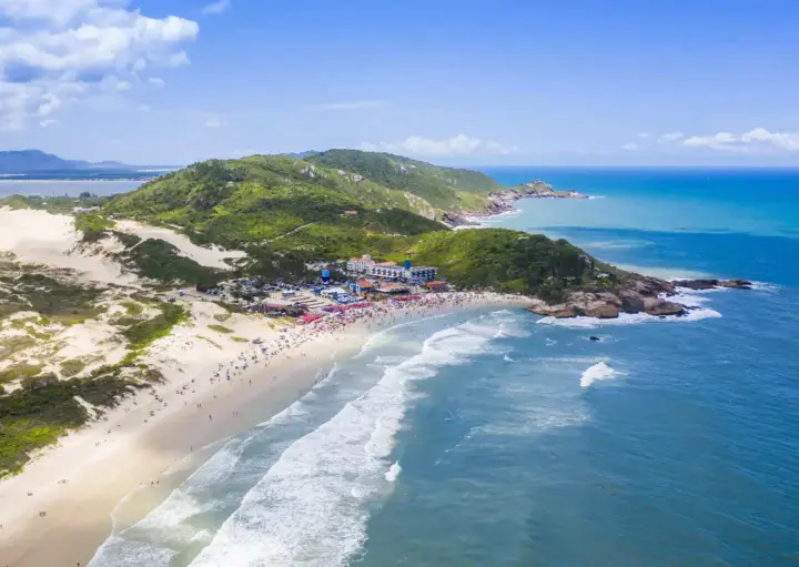 Imagem da vista aérea de uma praia de areia em Florianópolis cercada por ondas oceânicas azul-turquesa e colinas verdes. As pessoas se reúnem perto de edifícios coloridos ao longo da costa sob um céu azul brilhante com nuvens dispersas.