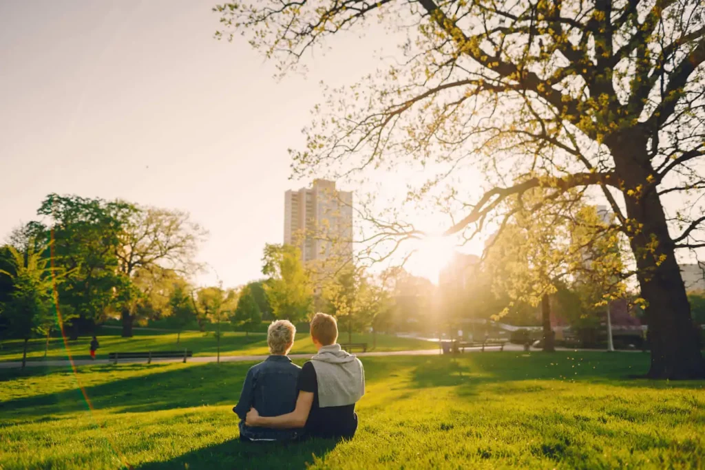 Imagem de duas pessoas sentadas juntas na grama de um parque, de costas e observando o pôr do sol. Árvores altas emolduram a cena, e um prédio da cidade é visível ao fundo sob um céu claro e dourado.
