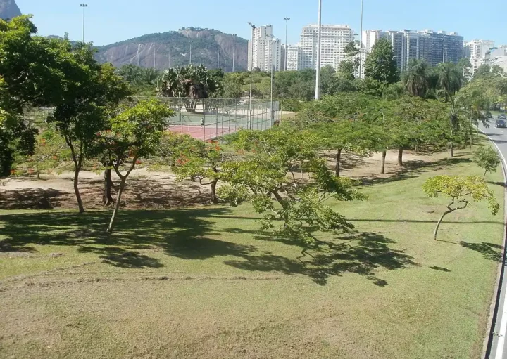 Um parque urbano com grama verde, árvores espalhadas e uma quadra de tênis, tendo como pano de fundo prédios altos e uma colina rochosa sob um céu azul claro.