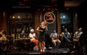 Uma mulher de vestido preto canta em um microfone no palco, cercada por músicos que tocam vários instrumentos em um animado local fechado com um letreiro de neon onde se lê Carioca da Gema atrás deles.