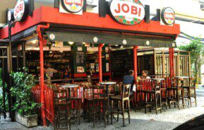 Vista externa de uma cafeteria vermelha e preta chamada Jobi, com mesas e cadeiras de madeira em um pátio de paralelepípedos; algumas pessoas estão sentadas, e os cardápios em quadro-negro são visíveis no interior. Plantas verdes emolduram a entrada.