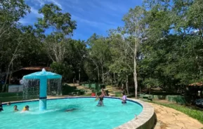 As crianças brincam em uma piscina externa circular com uma fonte azul central, cercada por árvores e vegetação sob um céu azul brilhante. Uma passarela de pedra corre ao longo da borda da piscina, com sapatos e garrafas colocados nas proximidades.