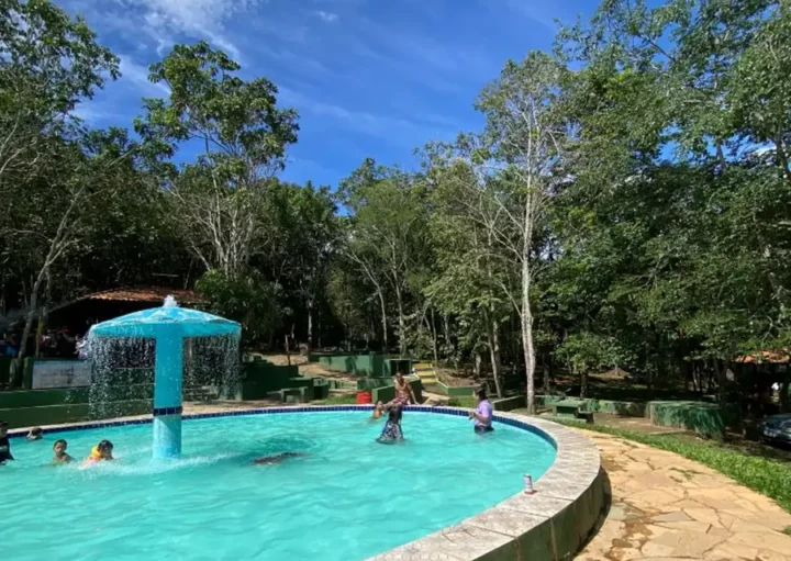 As crianças brincam em uma piscina externa circular com uma fonte azul central, cercada por árvores e vegetação sob um céu azul brilhante. Uma passarela de pedra corre ao longo da borda da piscina, com sapatos e garrafas colocados nas proximidades.