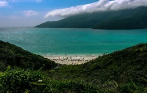 Vista de um oceano azul-turquesa cercado por colinas verdejantes, com uma praia de areia abaixo pontilhada de guarda-sóis e pessoas. Nuvens enevoadas pairam sobre montanhas distantes sob um céu azul brilhante.