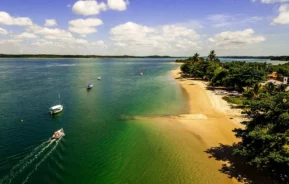 Vista aérea de uma praia tropical com areia dourada, água azul-esverdeada clara, alguns barcos e árvores exuberantes ao longo da costa sob um céu claro com nuvens dispersas.