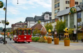 Um bonde vermelho viaja por uma rua da cidade repleta de prédios decorados, grandes esculturas de ovos de Páscoa e vasos de flores, criando uma atmosfera festiva. As pessoas caminham pelas calçadas ao fundo.