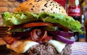 Um close-up de um hambúrguer gourmet com alface, tomate, picles, cebola roxa, bacon, queijo derretido e um hambúrguer de carne bovina em um pão com gergelim. Uma garrafa de Coca-Cola está borrada ao fundo.