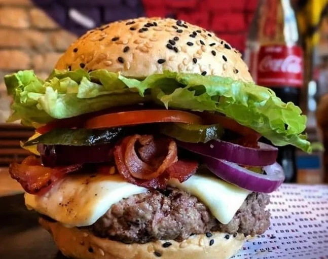 Um close-up de um hambúrguer gourmet com alface, tomate, picles, cebola roxa, bacon, queijo derretido e um hambúrguer de carne bovina em um pão com gergelim. Uma garrafa de Coca-Cola está borrada ao fundo.