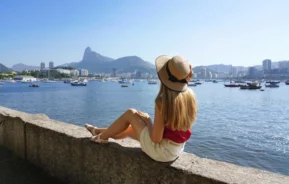 Uma mulher com um chapéu de sol senta-se na Mureta da Urca, observando os barcos na água com montanhas e prédios da cidade do Rio de Janeiro ao fundo em um dia claro e ensolarado.