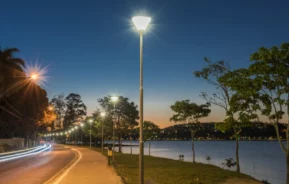 Uma estrada curva e arborizada passa ao lado de um lago calmo ao anoitecer, iluminada por postes de luz espaçados uniformemente. O céu passa do laranja próximo ao horizonte para o azul profundo acima, criando uma cena noturna tranquila.