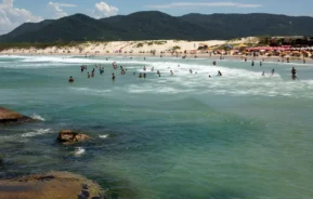 Pessoas nadando e brincando no oceano perto de uma praia de areia com guarda-sóis, com montanhas verdes e um céu azul ao fundo. Grandes rochas são visíveis na água azul-turquesa clara em primeiro plano.