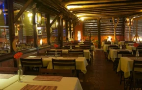 O interior de um restaurante aconchegante com vigas de madeira, toalhas de mesa amarelas em mesas quadradas e flores vermelhas simples em vasos de vidro. A iluminação quente e as grandes janelas criam uma atmosfera convidativa e rústica.