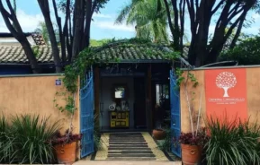 Entrada do restaurante Osteria Limoncello com portões azuis, paredes castanhas, vasos de plantas e uma placa com o logotipo de uma árvore branca. Árvores verdes e um telhado de telhas são visíveis ao fundo sob um céu azul.