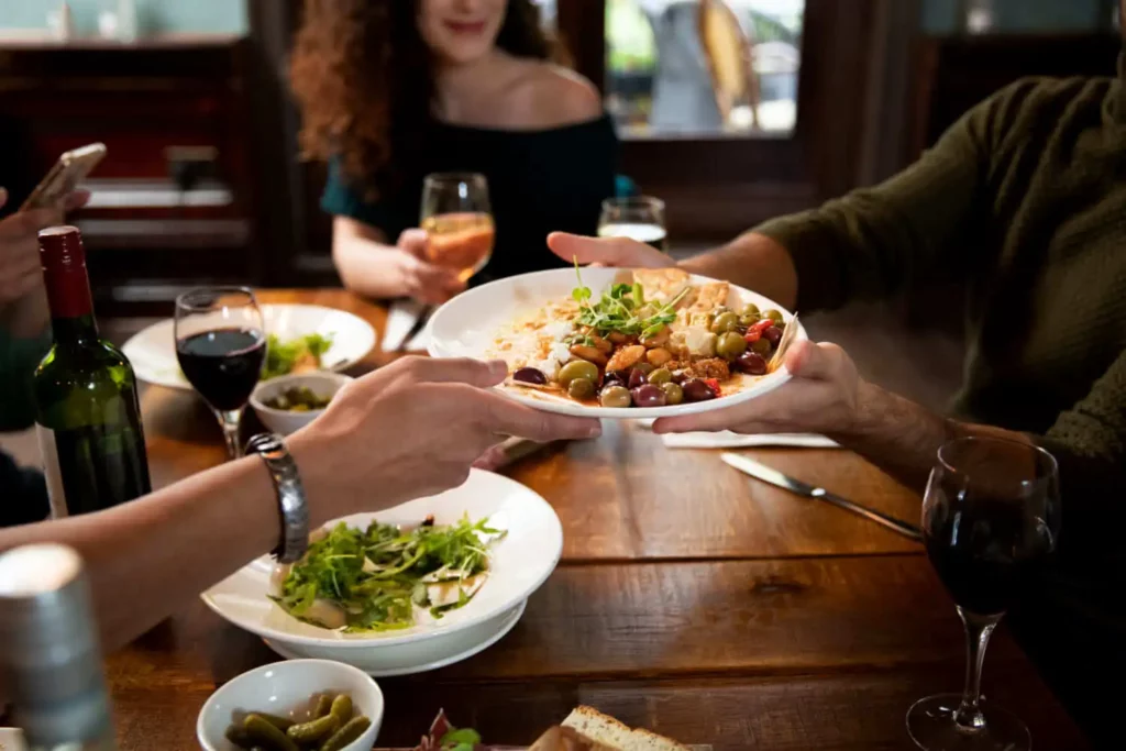 Imagem de pessoas compartilhando um prato de comida em uma mesa de madeira com taças de vinho, pratos de salada e pão. A atmosfera é casual e calorosa, sugerindo uma refeição social ou uma reunião entre amigos.
