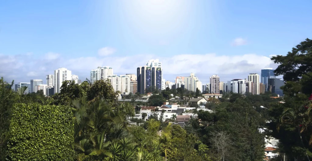Imagem da vista urbana de Alphaville, em Barueri, mostra uma paisagem ensolarada com altos edifícios modernos ao fundo e densas árvores verdes e casas residenciais em primeiro plano sob um céu azul claro