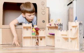 Imagem de um menino brincando com brinquedos de castelo de madeira e fantoches no chão, olhando para fora de uma estrutura de caixa de papelão que lembra uma casa, para ilustrar matéria sobre brinquedoteca de condomínio.